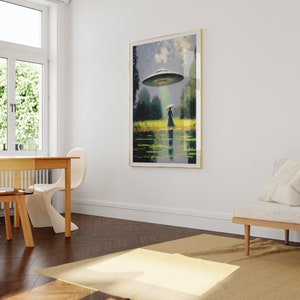 Op de afbeelding: Een ingelijst schilderij van een vrouw in een blauwe jurk die naar een vliegende schotel in een groen veld loopt. De schotel zweeft boven de vrouw en schijnt een lichtstraal op haar. Het schilderij hangt aan een witte muur in een kamer met een houten vloer.