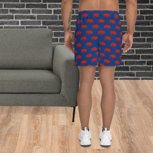 K&ouml;nnte beinhalten: Blaue Sporthose mit einem sich wiederholenden Muster aus roten B&uuml;ffelsilhouetten. Die Shorts bestehen aus leichtem Material und haben einen elastischen Bund. Die Person tr&auml;gt wei&szlig;e Turnschuhe.