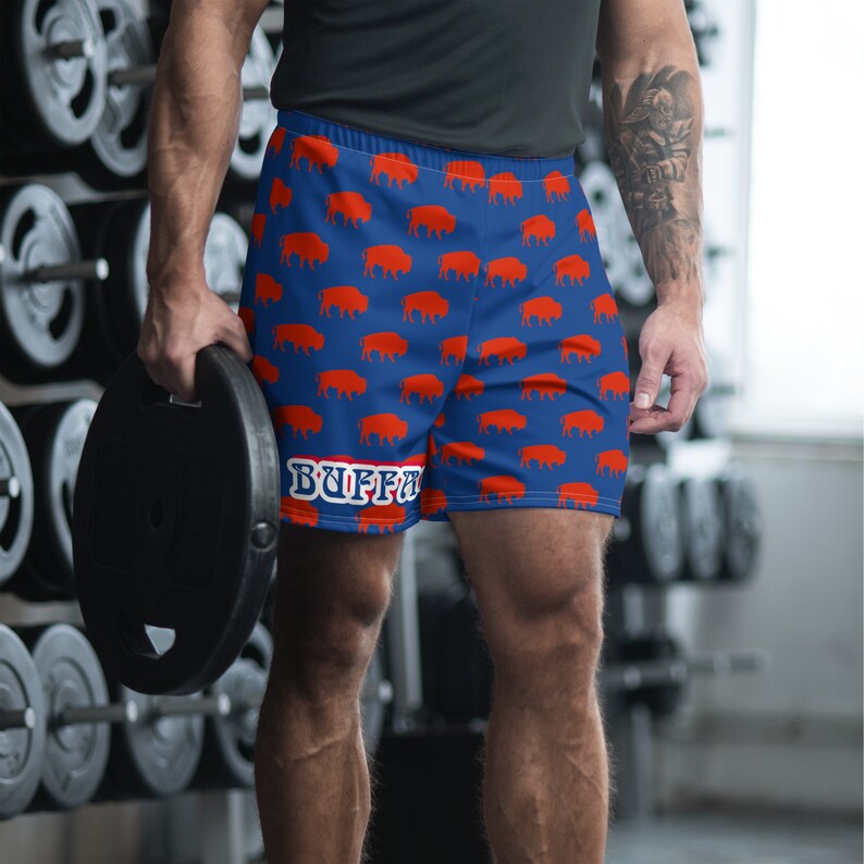 K&ouml;nnte beinhalten: Blaue Sporthosen mit einem sich wiederholenden Muster aus orangefarbenen B&uuml;ffelsilhouetten. Die Shorts haben einen wei&szlig;en Streifen und das Wort "BUFFALO" in wei&szlig;en Buchstaben. Hergestellt aus leichtem, atmungsaktivem Material.