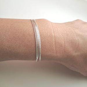 May include: A multi-strand silver bracelet consisting of five thin, polished metallic chains that wrap around the wrist. The bracelet features a sleek, uniform design with each individual strand maintaining a consistent, reflective silver finish.