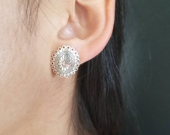 May include: Silver earrings with a circular design featuring a flower motif. The earrings are shown on a person's ear.