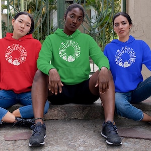 May include: Three people wearing hooded sweatshirts with a white graphic design of tools in a circle. The sweatshirts are red, green, and blue. The people are sitting on a concrete step.