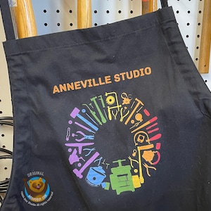 May include: Black apron with the text "ANNEVILLE STUDIO" in orange. A colorful circular design of tools is printed below the text. The apron is hanging on a pegboard with tools in the background.
