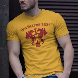 May include: Mustard yellow t-shirt with a red graphic featuring two dragons holding a hammer, the text "Twin Dragons Forge," and the year "1270." The shirt is short-sleeved and made of a soft material.