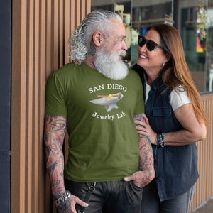 May include: A man and a woman standing in front of a store. The man is wearing a green t-shirt with the text "SAN DIEGO Jewelry Lab" and an image of an anvil and hammer. The woman is wearing a black vest over a white shirt.
