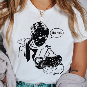Könnte beinhalten: Ein weißes T-Shirt mit einer schwarz-weißen Illustration eines Charakters, der eine Hockeymaske trägt und einen schwarzen Clog hält. Der Text "The hell?" befindet sich in einer Sprechblase über dem Charakter.