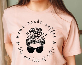 May include: A pink t-shirt with a black graphic design of a woman with a messy bun and sunglasses. The text on the shirt reads "mama needs coffee lots and lots of coffee".