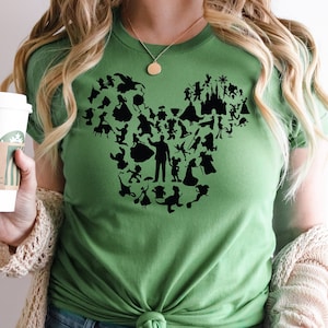 May include: Green t-shirt featuring a black silhouette of Mickey Mouse made up of Disney characters. The shirt is tied at the waist and the wearer is holding a coffee cup.