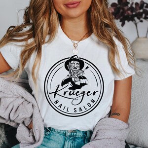 Op de afbeelding: Wit T-shirt met een zwart-witte afbeelding van Freddy Krueger die een schaar vasthoudt. De afbeelding bevindt zich in een cirkel met de tekst "Krueger Nail Salon".