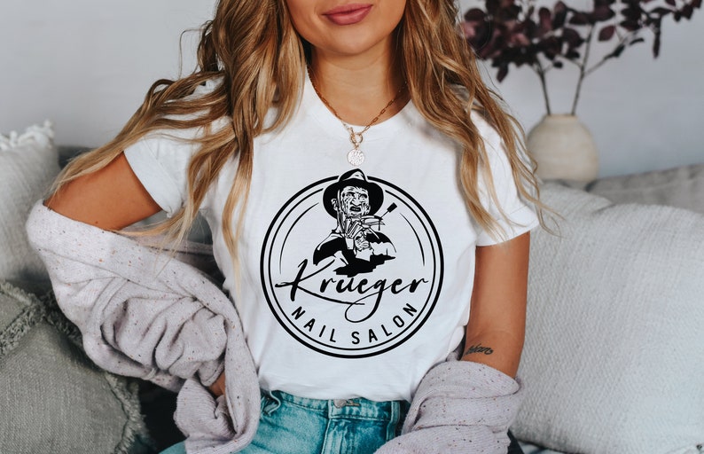 Op de afbeelding: Wit T-shirt met een zwart-witte afbeelding van Freddy Krueger die een schaar vasthoudt. De afbeelding bevindt zich in een cirkel met de tekst "Krueger Nail Salon".
