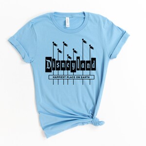 K&ouml;nnte beinhalten: Hellblaues T-Shirt mit einer schwarzen Grafik des Disneyland-Schilds mit Flaggen. Der Text "Happiest Place on Earth" befindet sich unter dem Schild.
