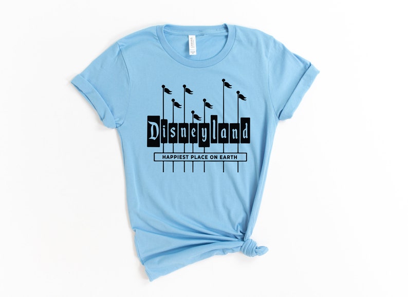 K&ouml;nnte beinhalten: Hellblaues T-Shirt mit einer schwarzen Grafik des Disneyland-Schilds mit Flaggen. Der Text "Happiest Place on Earth" befindet sich unter dem Schild.
