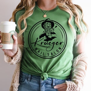 Op de afbeelding: Een groen T-shirt met een zwart-witte afbeelding van Freddy Krueger die nagelknippers vasthoudt. De afbeelding bevindt zich in een cirkel met de tekst "Krueger Nail Salon".