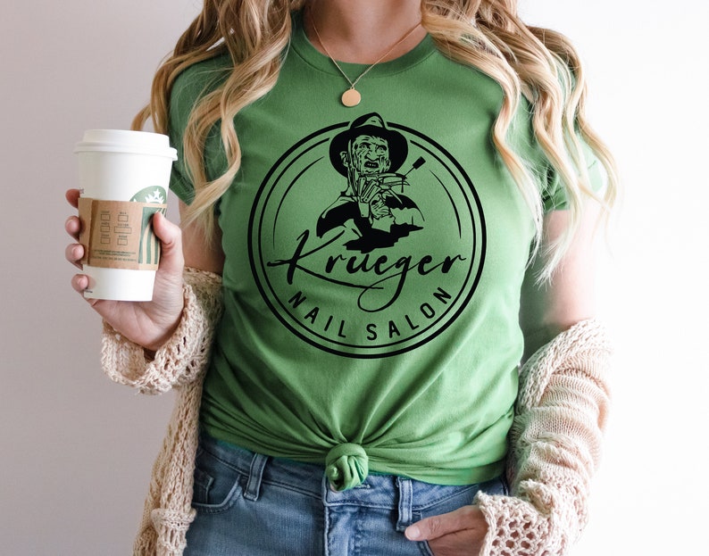 Op de afbeelding: Een groen T-shirt met een zwart-witte afbeelding van Freddy Krueger die nagelknippers vasthoudt. De afbeelding bevindt zich in een cirkel met de tekst "Krueger Nail Salon".