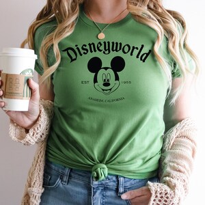K&ouml;nnte beinhalten: Gr&uuml;nes T-Shirt mit einem schwarz-wei&szlig;en Mickey Mouse-Motiv und dem Text "Disneyworld Est. 1955 Anaheim California".