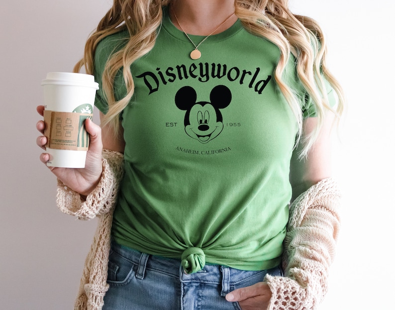 K&ouml;nnte beinhalten: Gr&uuml;nes T-Shirt mit einem schwarz-wei&szlig;en Mickey Mouse-Motiv und dem Text "Disneyworld Est. 1955 Anaheim California".