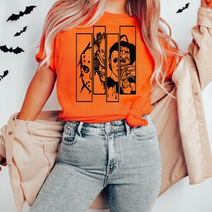 May include: A bright orange t-shirt with a black graphic of four horror movie characters. The characters are in black and white and are arranged in a row. The graphic includes Jason Voorhees, Michael Myers, Freddy Krueger, and Leatherface.