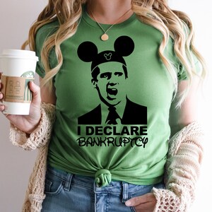 May include: A green t-shirt with a black and white image of a man wearing a Mickey Mouse hat. The text "I DECLARE BANKRUPTCY" is printed below the image.