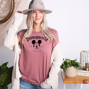 K&ouml;nnte beinhalten: Ein mauvefarbenes T-Shirt mit einem schwarzen Aufdruck von Mickey Mauses Kopf und dem Text "Disneyworld Est. 1955 Anaheim, Kalifornien".