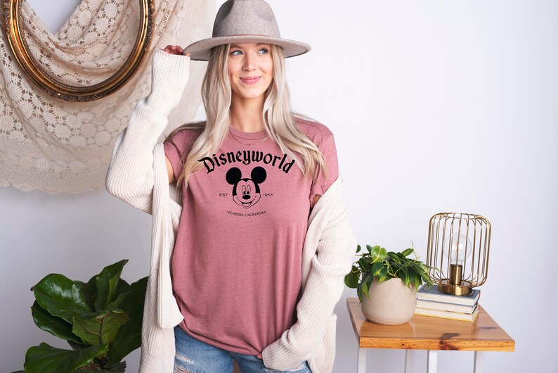 K&ouml;nnte beinhalten: Ein mauvefarbenes T-Shirt mit einem schwarzen Aufdruck von Mickey Mauses Kopf und dem Text "Disneyworld Est. 1955 Anaheim, Kalifornien".