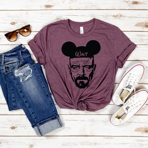 May include: A maroon t-shirt with a black graphic of Walter White from Breaking Bad wearing Mickey Mouse ears with the text "Walt" on the ear. The shirt is paired with blue jeans and white sneakers.