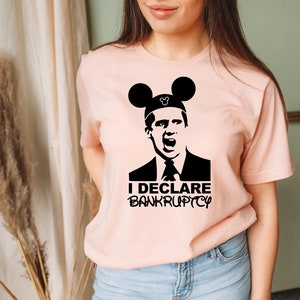 May include: A pink t-shirt with a black graphic of a character from the TV show "The Office" wearing Mickey Mouse ears. The text "I DECLARE BANKRUPTCY" is printed below the character.