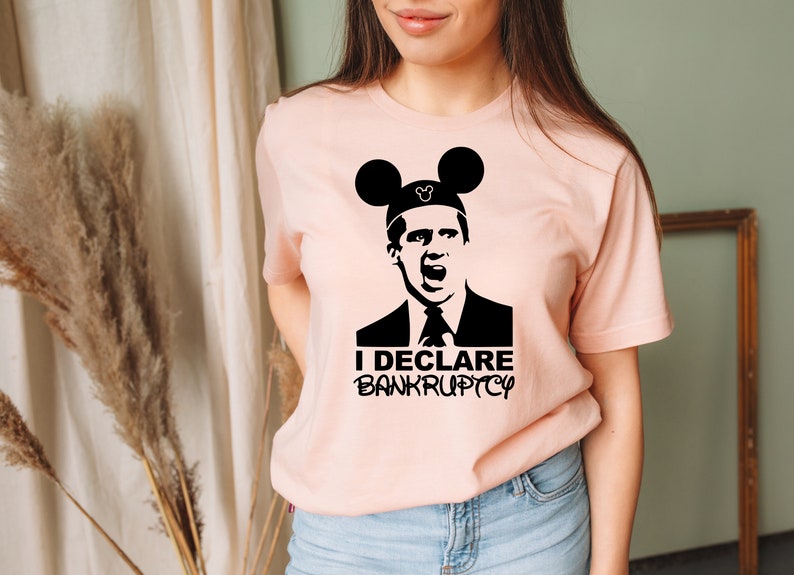 May include: A pink t-shirt with a black graphic of a character from the TV show "The Office" wearing Mickey Mouse ears. The text "I DECLARE BANKRUPTCY" is printed below the character.