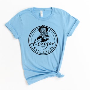 Op de afbeelding: Lichtblauw T-shirt met een zwart-wit logo met de tekst "Krueger Nail Salon". Het logo toont een cartoonfiguur dat een vijl vasthoudt.