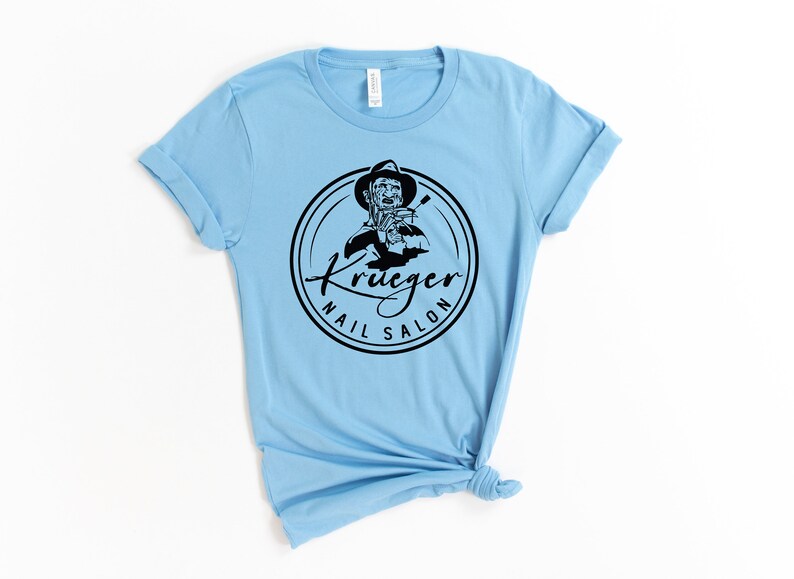 Op de afbeelding: Lichtblauw T-shirt met een zwart-wit logo met de tekst "Krueger Nail Salon". Het logo toont een cartoonfiguur dat een vijl vasthoudt.