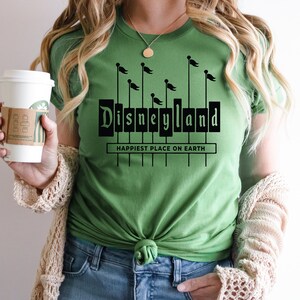 K&ouml;nnte beinhalten: Ein gr&uuml;nes T-Shirt mit einer schwarzen Grafik des Disneyland-Schilds und dem Text "Happiest Place on Earth".