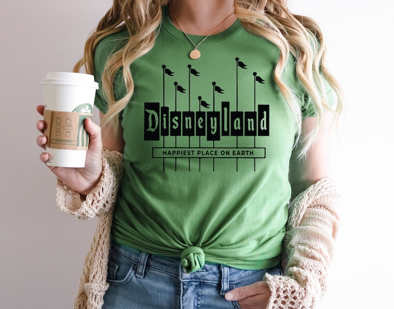K&ouml;nnte beinhalten: Ein gr&uuml;nes T-Shirt mit einer schwarzen Grafik des Disneyland-Schilds und dem Text "Happiest Place on Earth".