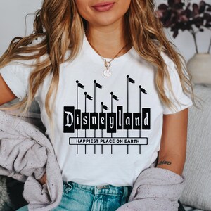K&ouml;nnte beinhalten: Ein wei&szlig;es T-Shirt mit einer schwarzen Grafik des Disneyland-Schilds mit Flaggen und dem Text "Happiest Place on Earth".