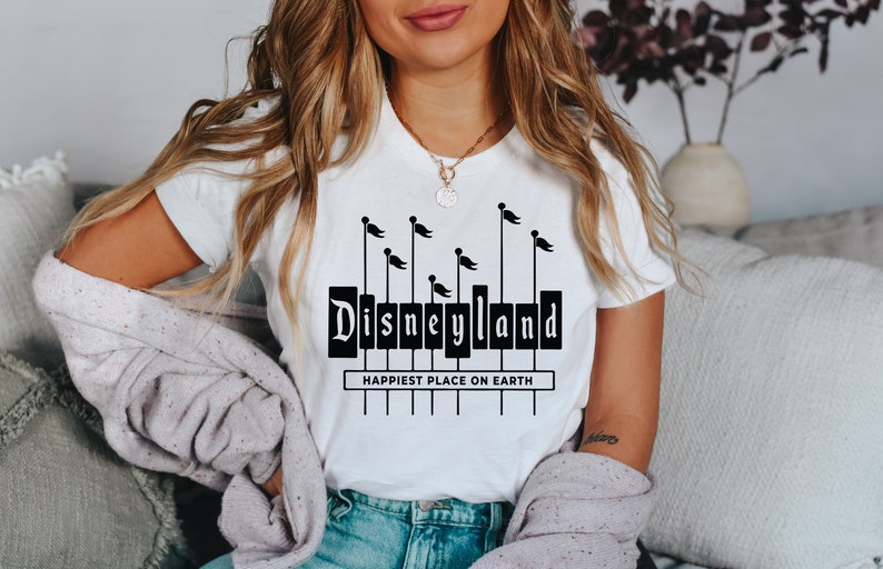 K&ouml;nnte beinhalten: Ein wei&szlig;es T-Shirt mit einer schwarzen Grafik des Disneyland-Schilds mit Flaggen und dem Text "Happiest Place on Earth".