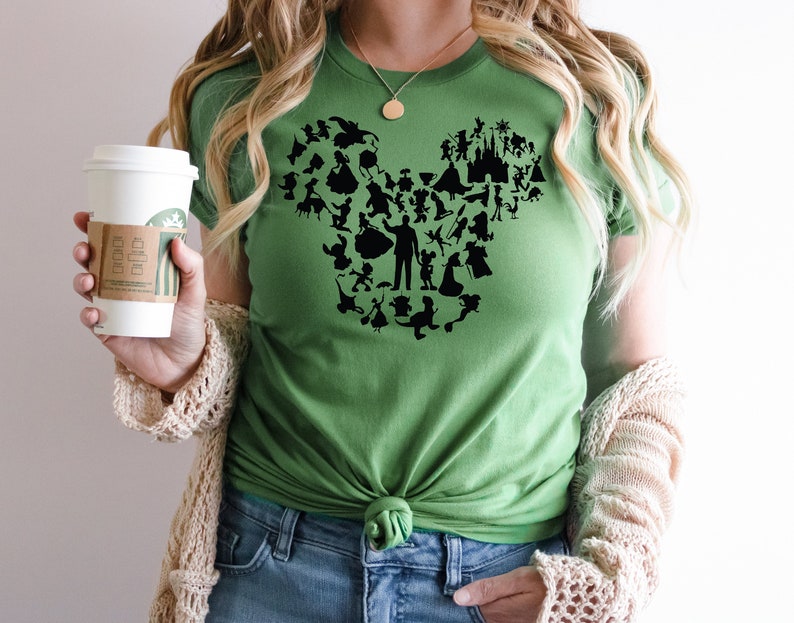 May include: Green t-shirt featuring a black silhouette of Mickey Mouse made up of Disney characters. The shirt is tied at the waist and the wearer is holding a coffee cup.
