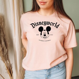 K&ouml;nnte beinhalten: Ein rosa T-Shirt mit einem schwarzen Aufdruck von Mickey Mouse und dem Text "Disneyworld Est. 1955 Anaheim, Kalifornien".