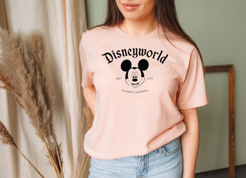 K&ouml;nnte beinhalten: Ein rosa T-Shirt mit einem schwarzen Aufdruck von Mickey Mouse und dem Text "Disneyworld Est. 1955 Anaheim, Kalifornien".