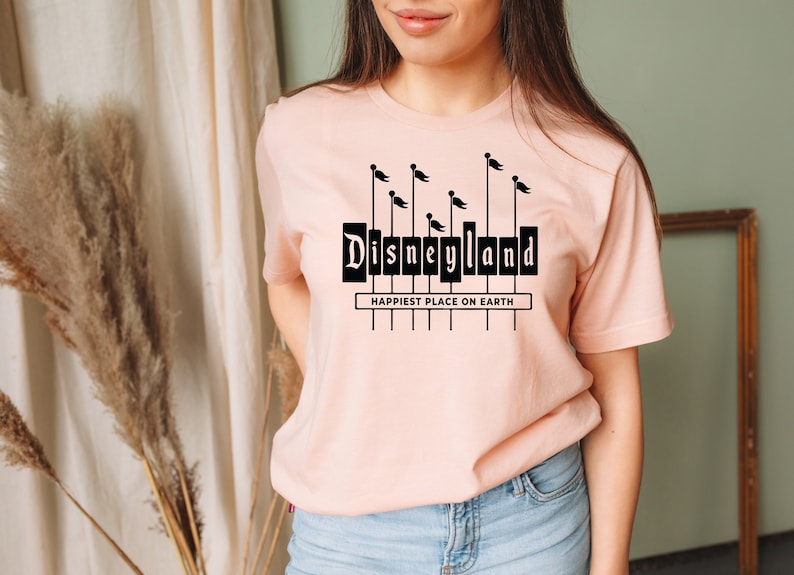 K&ouml;nnte beinhalten: Ein rosa T-Shirt mit einer schwarzen Grafik des Disneyland-Schilds und dem Text "Happiest Place on Earth".