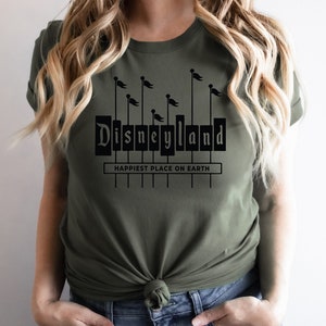 K&ouml;nnte beinhalten: Ein gr&uuml;nes T-Shirt mit einer schwarzen Grafik des Disneyland-Schilds mit dem Text "Happiest Place on Earth" darunter.