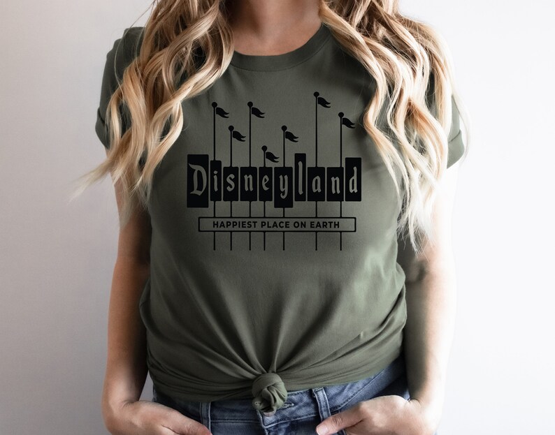 K&ouml;nnte beinhalten: Ein gr&uuml;nes T-Shirt mit einer schwarzen Grafik des Disneyland-Schilds mit dem Text "Happiest Place on Earth" darunter.