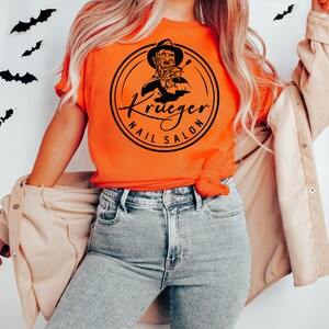 Op de afbeelding: Een vrouw draagt een oranje T-shirt met een zwart-witte afbeelding van Freddy Krueger die nagelknippers vasthoudt en de tekst "Krueger Nail Salon".