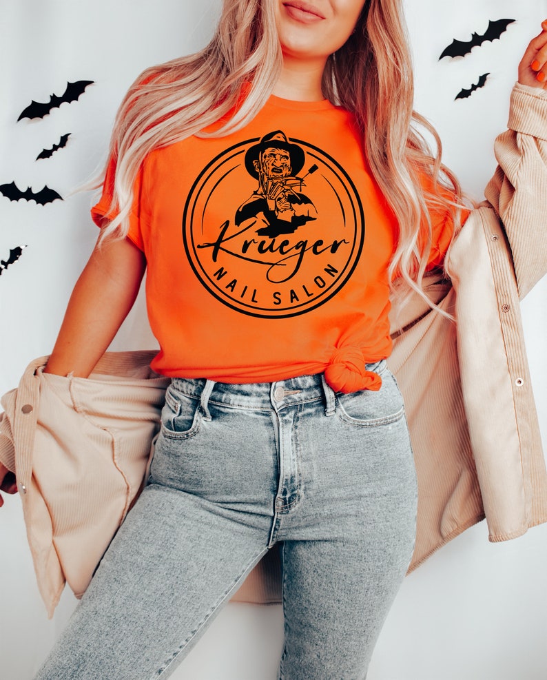 Op de afbeelding: Een vrouw draagt een oranje T-shirt met een zwart-witte afbeelding van Freddy Krueger die nagelknippers vasthoudt en de tekst "Krueger Nail Salon".