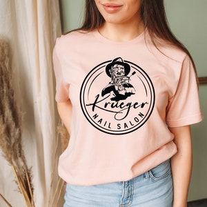 Op de afbeelding: Een roze T-shirt met een zwart-witte afbeelding van een persoon die een nagelvijl vasthoudt. De afbeelding bevindt zich in een cirkel met de tekst "Krueger Nail Salon".