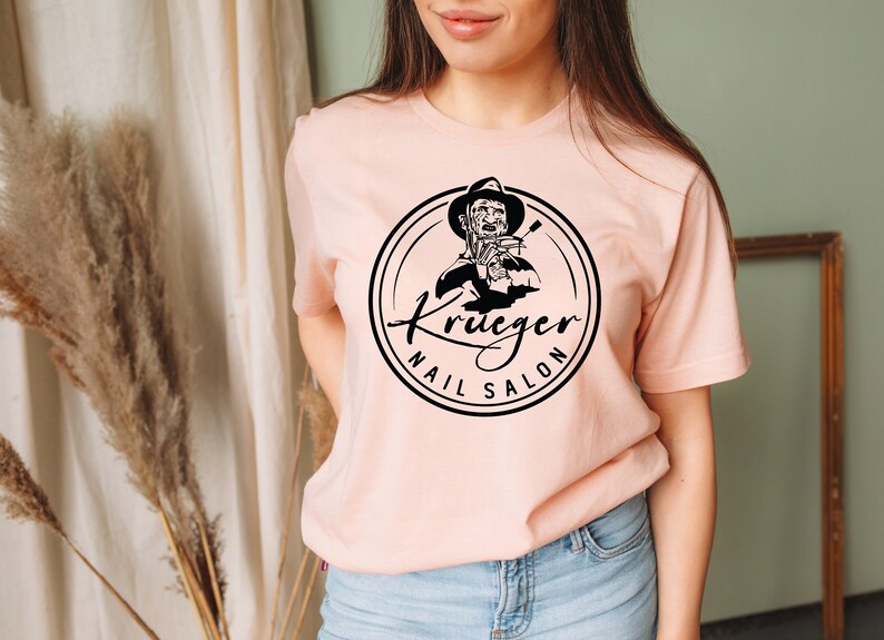 Op de afbeelding: Een roze T-shirt met een zwart-witte afbeelding van een persoon die een nagelvijl vasthoudt. De afbeelding bevindt zich in een cirkel met de tekst "Krueger Nail Salon".
