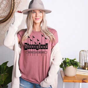 K&ouml;nnte beinhalten: Ein pinkfarbenes T-Shirt mit einer schwarzen Grafik des Disneyland-Schilds mit dem Text "Happiest Place on Earth".