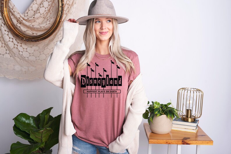 K&ouml;nnte beinhalten: Ein pinkfarbenes T-Shirt mit einer schwarzen Grafik des Disneyland-Schilds mit dem Text "Happiest Place on Earth".