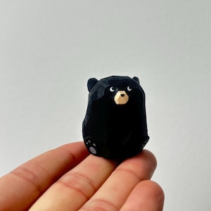 Könnte beinhalten: Kleine, schwarze Bärenfigur mit geschnitztem Aussehen. Der Bär hat eine hellbraune Schnauze und kleine, weiße Augen. Die Figur wird in der Hand einer Person vor einem schlichten weißen Hintergrund gehalten.