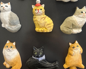 May include: A collection of nine carved wooden cat figurines in various colors and poses. The cats range from gray to orange and black, with some wearing hats or bows. The figurines are displayed against a dark background.