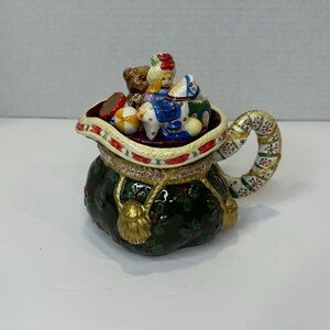 Peut inclure: Une crémier en céramique verte en forme de sac de Père Noël avec un pompon doré et des accents de houx. La crémier est remplie de jouets miniatures en céramique, dont un ours en peluche, une poupée, un cheval à bascule et un ballon.