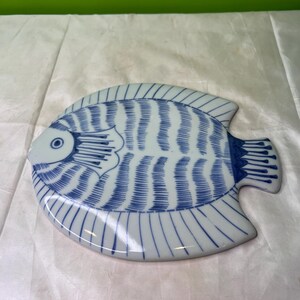 May include: A ceramic fish-shaped plate with a blue and white design. The plate features a detailed illustration of a fish with blue stripes and a white background. The plate is approximately 25 cm long.