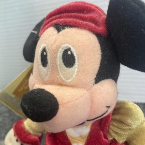 May include: A plush Mickey Mouse character toy featuring a peach-colored face, large black ears, and a red velvet-textured hat. The character wears a red velvet vest over gold sleeves, with embroidered eyes and a black nose, capturing a classic look.